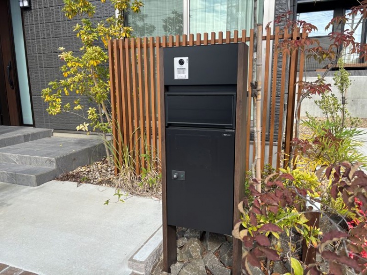 ダークグレーの外壁の２階建て住宅のB棟。玄関前に設置された黒い宅配ボックスと周囲には植栽が植えられています。