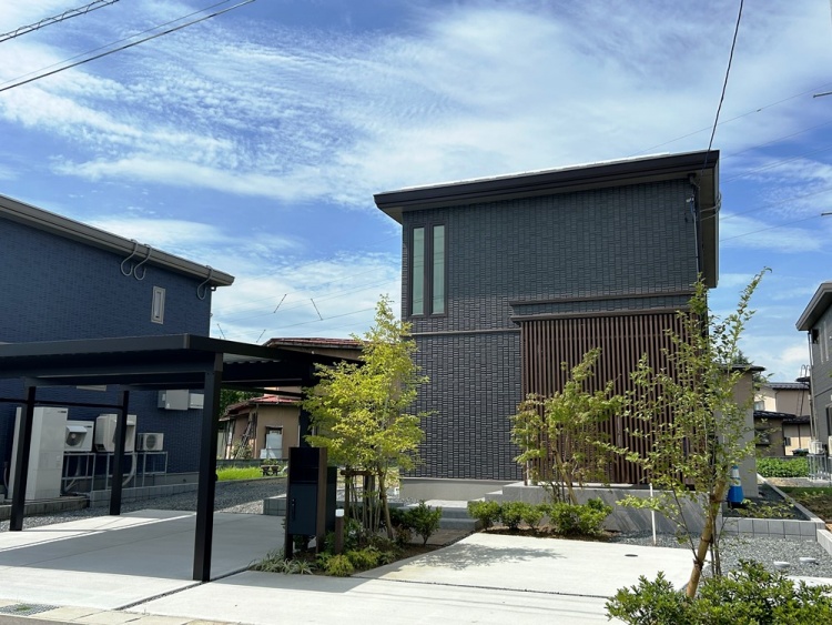 ダークグレーを基調とした外壁を持つ2階建て住宅の外観。前にはカーポートや緑豊かな庭が前景に広がっています。空には青空が広がり、静かな住宅街の一角が描かれています。