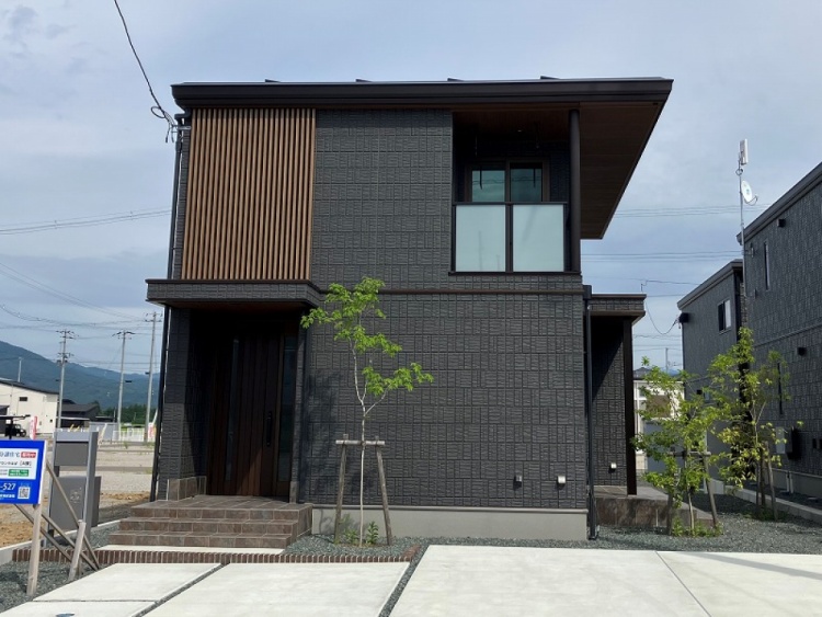 ダークグレーの外壁を持つ2階建ての住宅が、夕暮れの下に建っています。前には小さな庭があり、若木が植えられています。