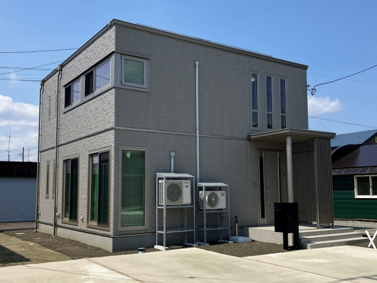 グレーを基調とした二階建て住宅の外観です。縦長の窓と大きなガラス窓が特徴で、玄関にはデザインパネルが設置されています。外壁には2台の空調設備が取り付けられており、玄関には数段の階段が続いています。