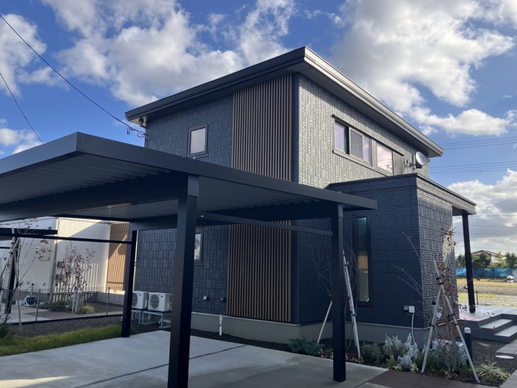 グレーの外壁と木目のアクセントが特徴的な2階建ての家が、青空の下にあります。家の前には広々とした駐車場とカーポートがあり、周囲には緑豊かな植栽が施されています。