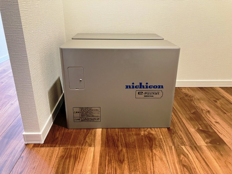 床に設置されたグレーの蓄電池で、壁に沿って設置されています。機器には「nichicon」と青い文字が書かれており、コードが壁に繋がっています。