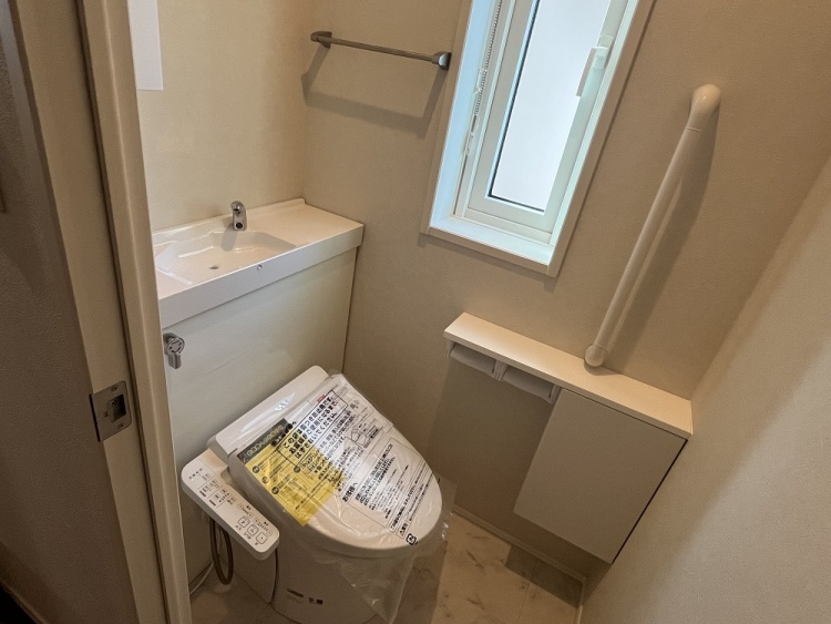 コンパクトながら機能的なトイレ空間。白を基調とした内装に、小窓からの自然光が心地よく差し込む。手洗い器や収納棚、手すりが備わり、使い勝手の良い設計になっている。