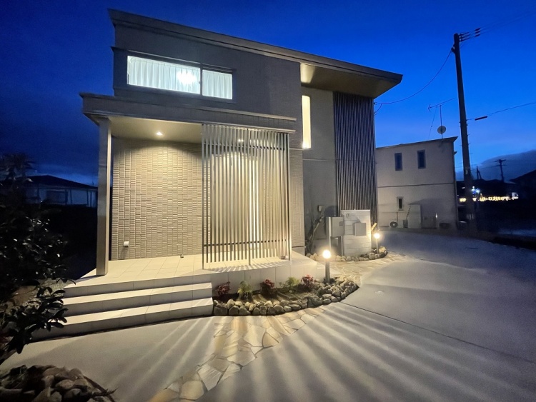 グレーの外壁が特徴的な2階建て住宅の外観。玄関前には縦格子の目隠しがあり、階段を上がった先に玄関があります。庭には低木や石が配置され、外観はすっきりとした印象です。