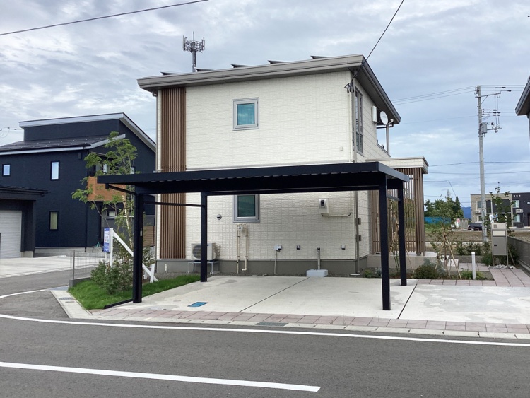 淡いグレーの外壁に木目のアクセントが映える2階建ての家が、角地に建っています。家の前にはカーポートがあり、植栽が施された庭が周囲を彩っています。