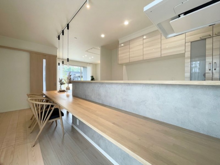 キッチンカウンターに沿って長い木製のテーブルがあり、椅子が並んでいます。柔らかな光が差し込み、リラックスした雰囲気のダイニングスペースです。