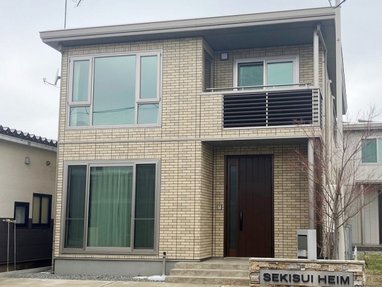 落ち着いたベージュの外壁が特徴的な二階建ての住宅。大きな窓が開放感を生み出し、バルコニーの黒い手すりがアクセントに。玄関までのアプローチには石畳が敷かれ、シンプルながらも上品な雰囲気。紅葉した木が庭に彩りを添えている。
