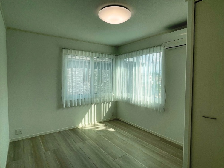 明るい窓が2面にあるシンプルな洋室。天井には丸い照明があり、白いカーテン越しに自然光が差し込んでいます。木目調の床が温かい雰囲気を演出しています。