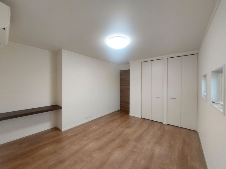 木目の床と白い壁が広がる部屋で、窓から柔らかな光が差し込み、奥の壁には小さな棚があります。