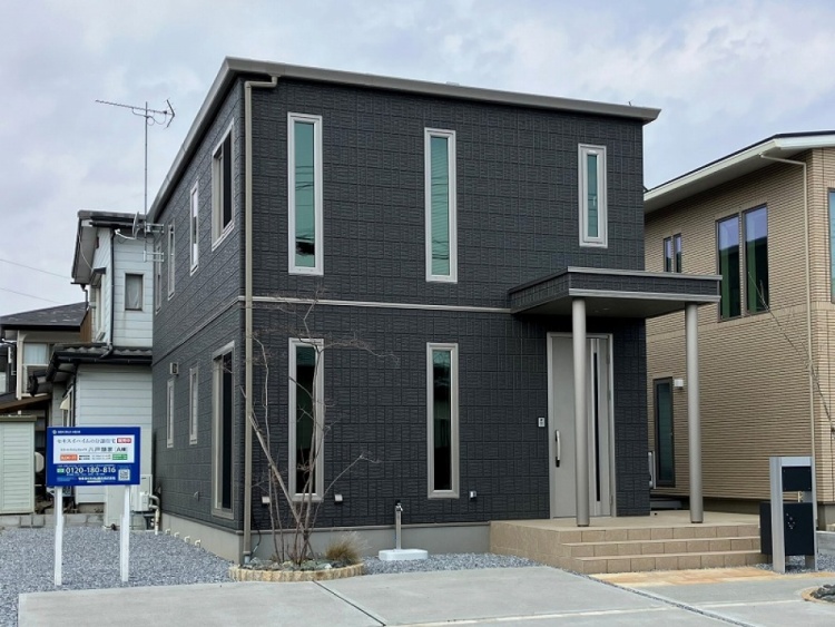 ダークグレーを基調とした二階建て住宅の外観で、縦長の窓がいくつも並んでいます。玄関はシルバーのドアが設置され、小さなポーチがあるモダンなデザインです。前庭には若い木が植えられており、整った外観が印象的です。