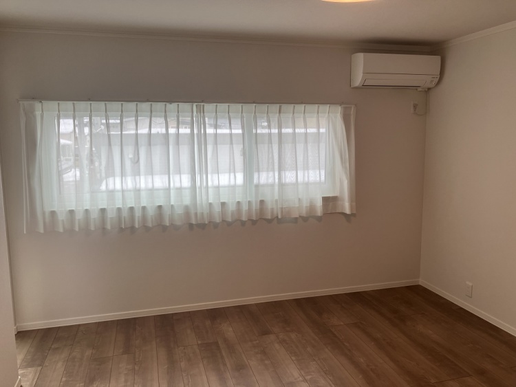 木目調の床が温かみを感じさせるシンプルな部屋。横長の大きな窓には白いカーテンがかかり、優しい光が室内を包みます。壁にはエアコンが設置され、快適な空間になっています。