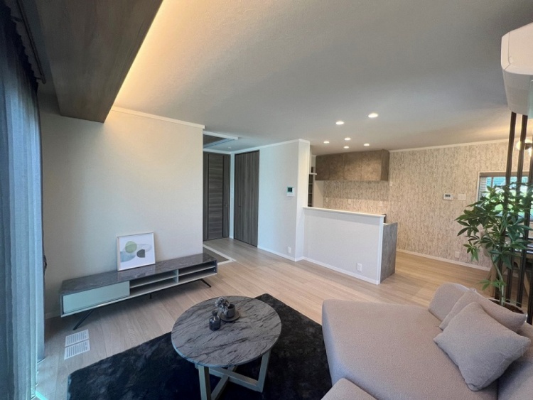 リビングには丸い大理石模様のテーブルとベージュのソファがあり、黒いラグが敷かれています。左手にはアートが飾られたテレビ台があり、奥にはキッチンカウンターが見えます。