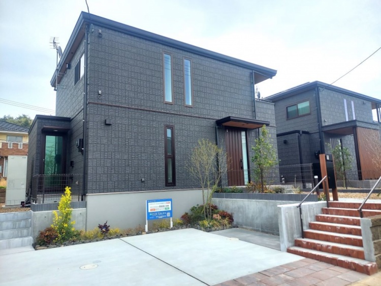 シックな外壁と縦長の窓が印象的な二階建て住宅。植栽が玄関まわりを彩り、自然と調和した落ち着いた雰囲気を演出している。