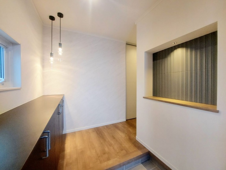 玄関の正面に吊り下げられた照明が柔らかく輝き、黒い収納と木の床が落ち着いた雰囲気をつくっています。