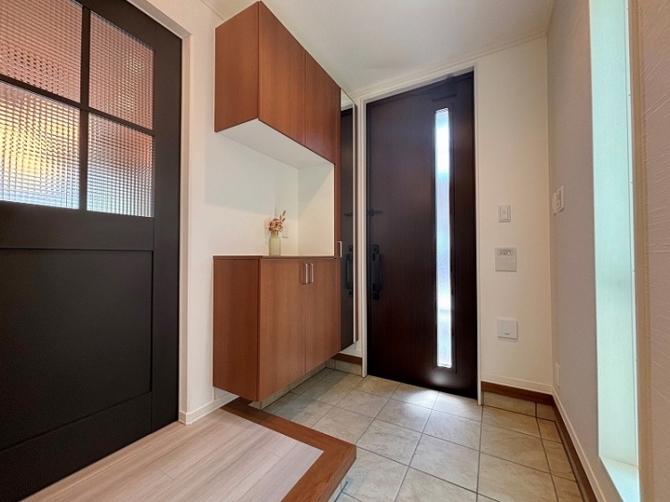 玄関エリアの写真です。床はタイル張りで、右側にスリムな縦長の窓が付いた黒い玄関ドアがあります。左側にはガラスの入った黒いドアがあり、正面には木製の収納棚が設置されています。棚の上には小さな花瓶が飾られ、温かみのある雰囲気です。