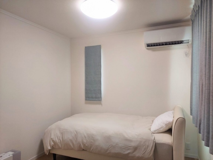 シンプルで落ち着いた雰囲気のベッドルーム。白い壁と明るい木目調の床に、ベージュのベッドが設置されています。