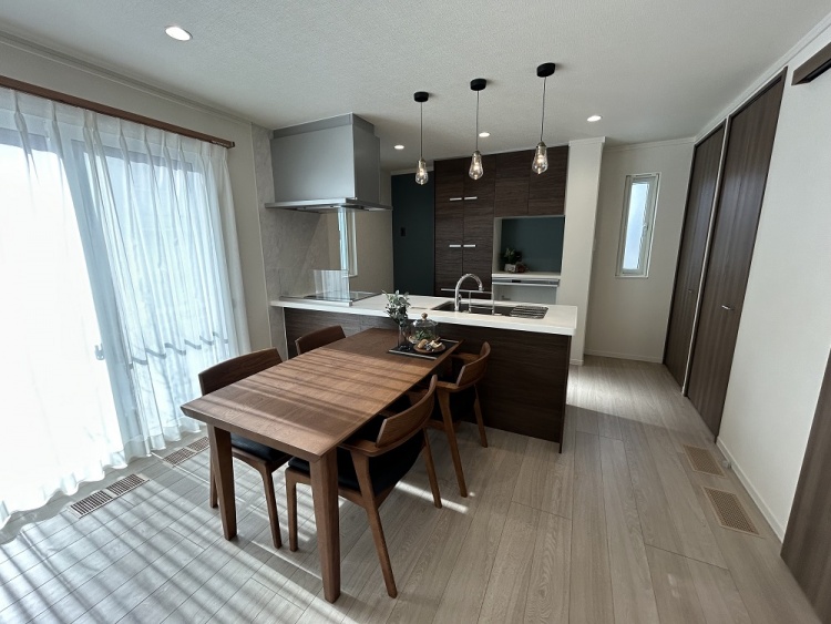 明るい光が差し込むダイニングキッチンの風景。木目のダイニングテーブルには4脚の椅子が並び、中央には小さな植物と食器が置かれている。奥には白い天板のキッチンカウンターがあり、黒い吊り下げ照明が洗練された雰囲気を演出している。収納が豊富なキッチンキャビネットが並び、落ち着いた木のぬくもりを感じる空間となっている。