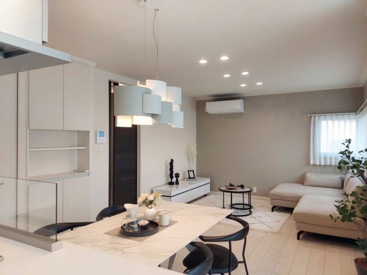 大理石調のテーブルと黒い椅子が置かれたダイニングエリア。奥にはL字型の白いソファと観葉植物が置かれたリビングがあり、グレーの壁と白い床が空間全体に明るく洗練された印象を与えている。