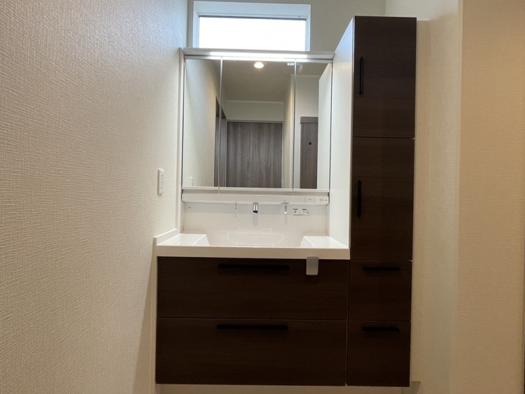 三面鏡付きの洗面化粧台です。ミラー下に一体型の洗面ボウルと引き出し収納があり、右側には縦型の収納キャビネットを備えた、使い勝手の良い洗面スペースです。