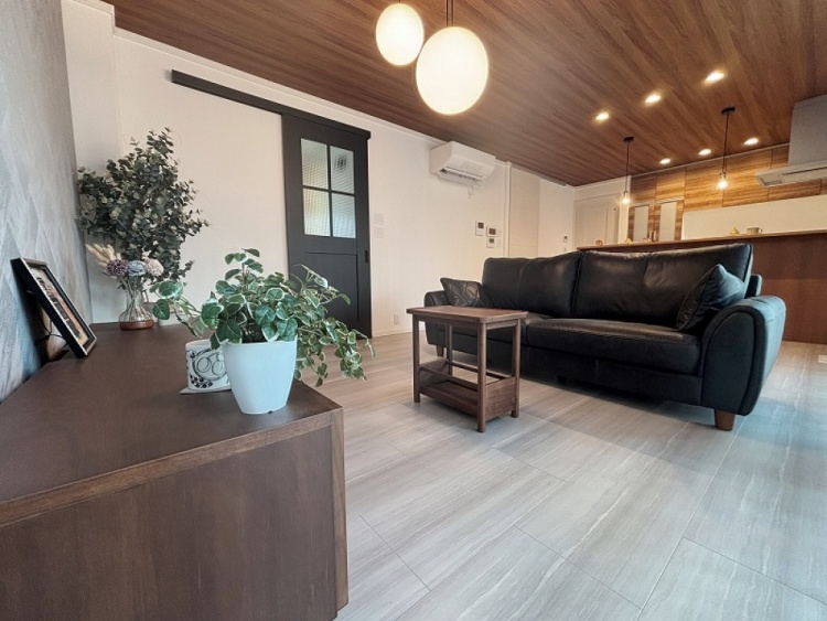 リビングルームの写真です。中央には黒い革製のソファがあり、手前からその背もたれが見えています。壁際には木製のテレビ台が置かれ、上に小物や写真立てが飾られています。右側には観葉植物が置かれており、黒い引き戸が部屋のアクセントになっています。天井からは2つの丸いペンダントライトが下がり、部屋全体に柔らかな光を放っています。天井は木目調で、温かみのある空間を演出しています。