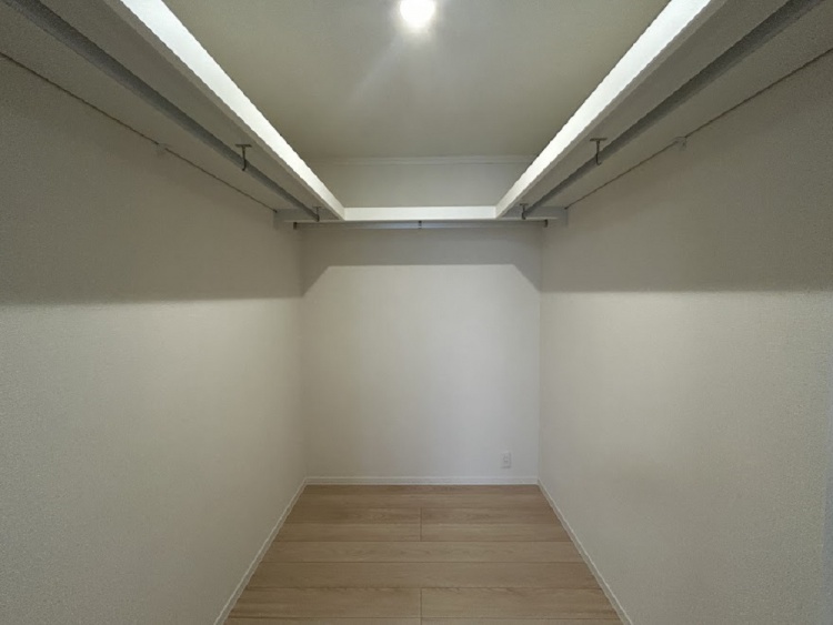 白い壁と木目の床が広がるシンプルな部屋。奥にはクローゼットの扉があり、左側の窓から優しい光が差し込んでいる。温かみのある落ち着いた空間。