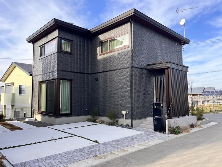 黒い外壁が印象的な二階建ての家が静かな住宅街に建っており、手前には白い駐車スペースと石畳のアプローチが広がっています。