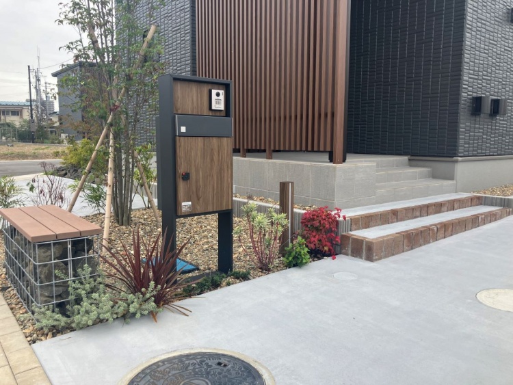 黒い外壁と木の格子が印象的な二階建ての家で、玄関前には緑が彩りを添えています。