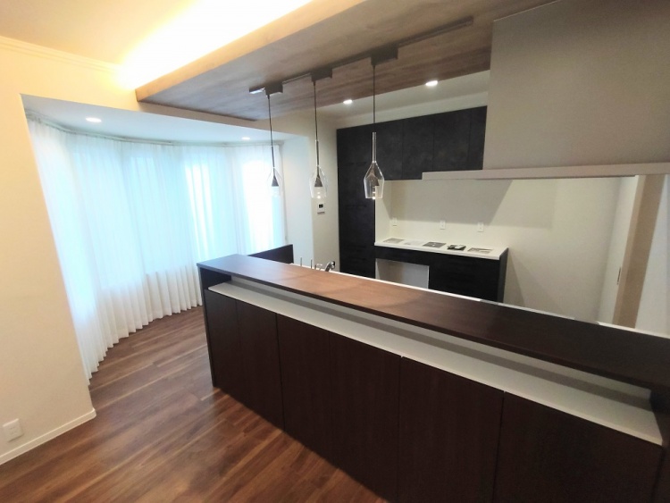 広々としたリビングダイニング。木目の美しい床と、ダークカラーのキッチンが落ち着いた雰囲気を作る。右手には階段があり、奥にはペンダントライトが揺れるキッチンカウンターが見える。