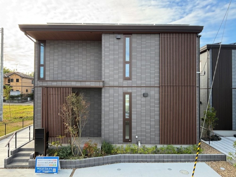 青空の下、濃い色の外壁と木の格子が調和した二階建ての家が堂々と立っています。