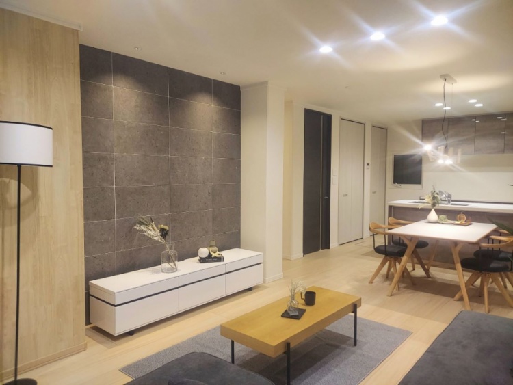 明るいリビングダイニングの風景。木製のダイニングテーブルと椅子が中央にあり、奥には対面式のキッチンが見える。右側には観葉植物とグレーのソファが置かれ、窓からの日差しが温かみを添えている。