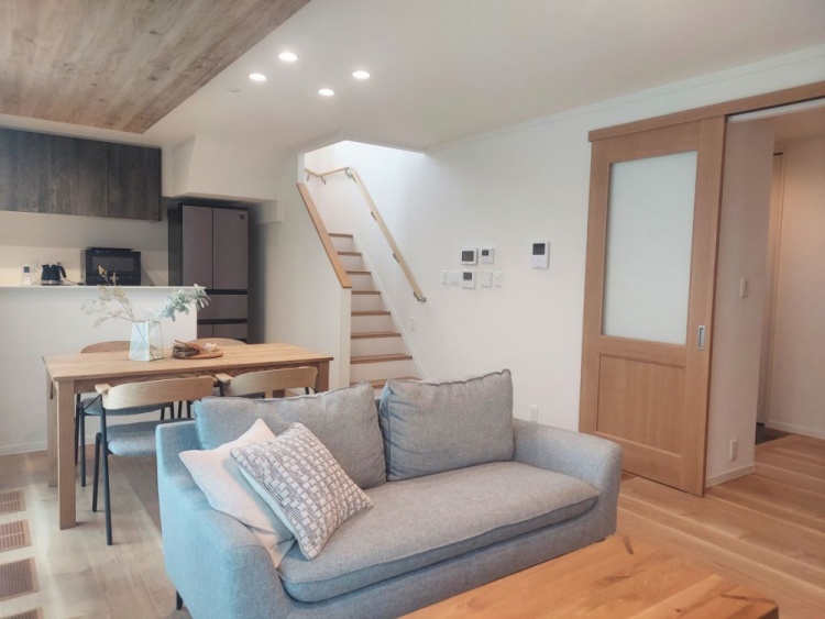 淡い木目の床と天井に包まれた明るいリビングダイニング。グレーのソファと木製のテーブルがあり、奥には白い階段が続いています。