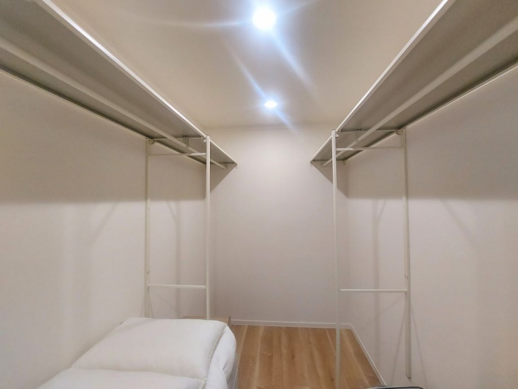 シンプルで広々としたウォークインクローゼット。両側にパイプハンガーと棚が設置されており、衣類や収納ボックスを効率的に整理可能。天井にはダウンライトがあり、明るく見通しの良い空間です。