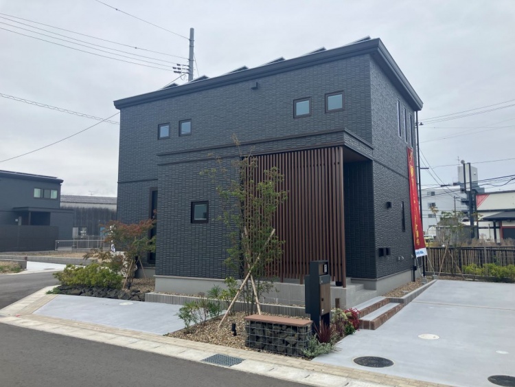 ダークグレーの外壁と縦格子の玄関まわりが印象的な、モダンな2階建て住宅。広い駐車スペースとシンプルな植栽が整備された外構デザイン。