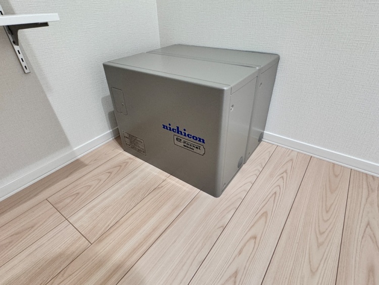 床に設置されたグレーの蓄電池で、壁際に置かれています。機器には「nichicon」と青いロゴがあり、床は明るい木目調です。