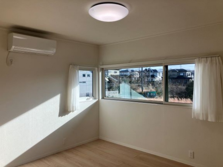 大きな窓からたっぷりと日差しが差し込む明るい部屋。外には住宅や木々が見え、穏やかな景色が広がっている。壁にはエアコンが設置され、快適な空間が整えられている。
