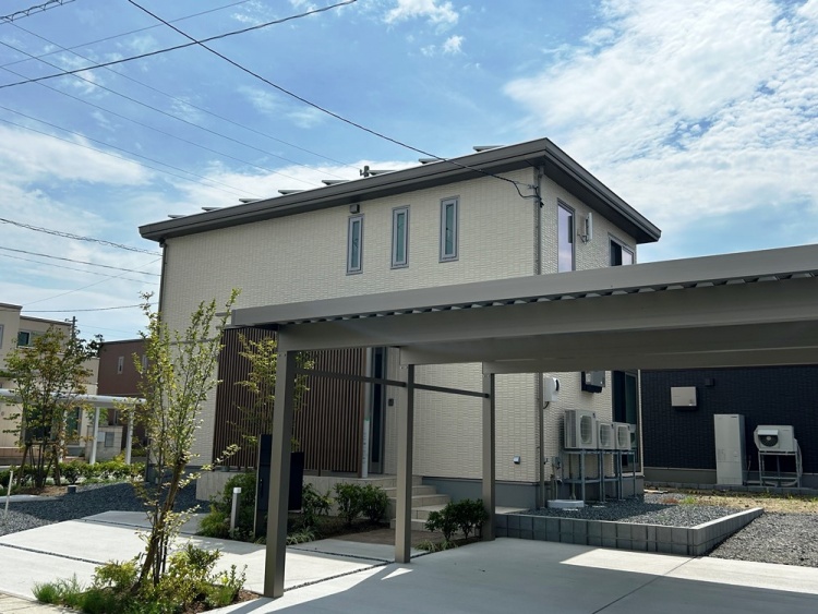 ベージュを基調とした外壁の2階建て住宅が、青空の下に建っています。前には広いカーポートがあり、植栽や小さな庭が整えられています。