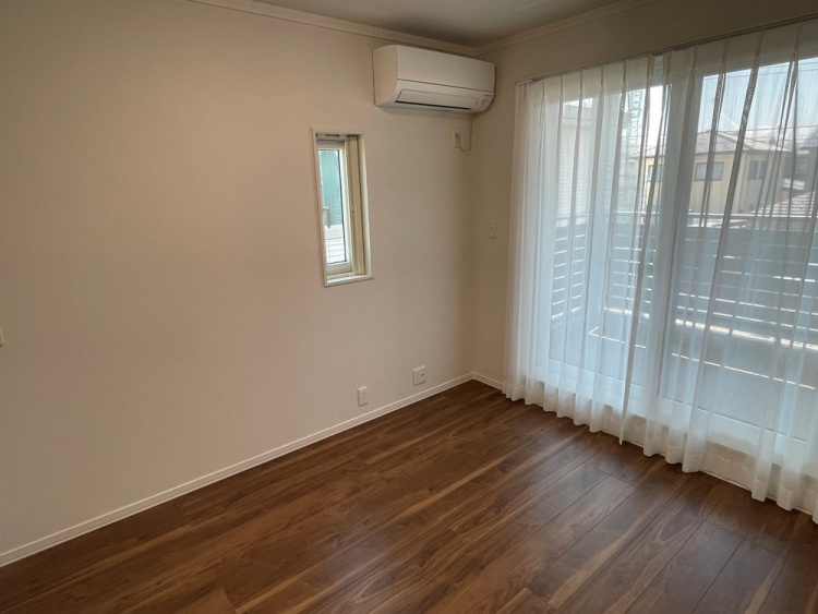 木目調の床が広がる明るい部屋です。大きな窓には白いカーテンがかかっており、外の光が優しく差し込んでいます。左側にはエアコンが設置されており、奥には白い引き戸が見えます。