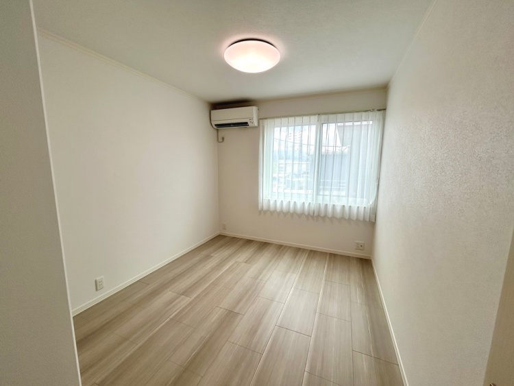 大きな窓から光が差し込むシンプルな部屋。白いカーテンとエアコンが備え付けられ、天井には丸い照明が優しい光を放っています。木目調の床が部屋全体に温かみを与えています。