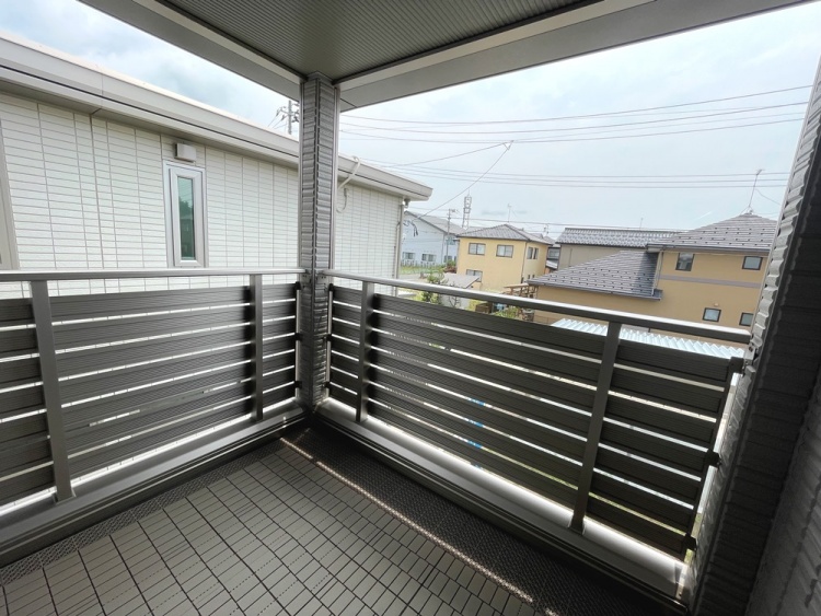 ‘屋根付きのバルコニーで、金属製の手すりに囲まれています。床はグレーのタイルで、周囲には住宅が並んでおり、青空が広がっています。