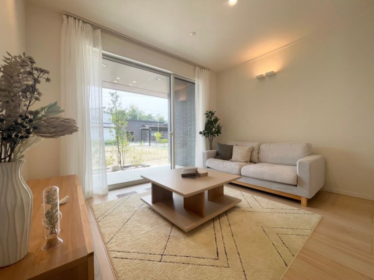 大きな窓から庭が見える明るいリビングルーム。白いソファと木製のテーブルが配置され、柔らかな光が部屋全体に広がっています。