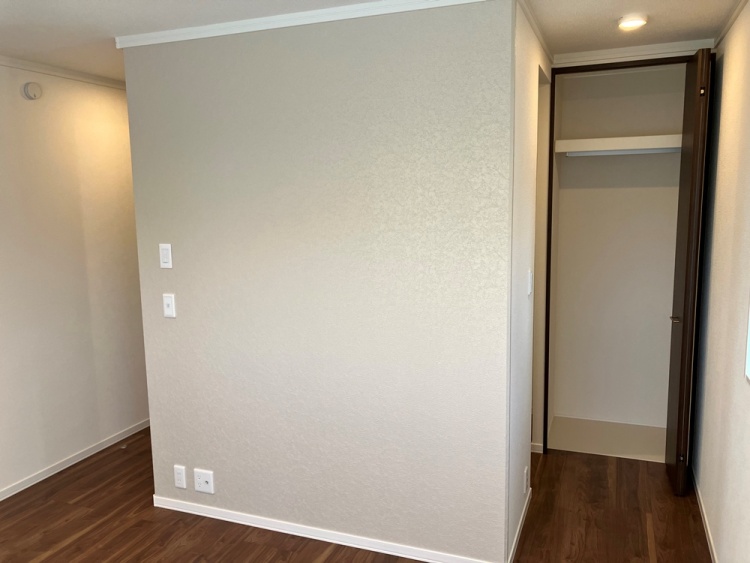 木目調の床に白い壁が調和する主寝室の一角に、クローゼットの扉が開かれています。天井には照明が設置され、壁にはスイッチとコンセントが配置されています。