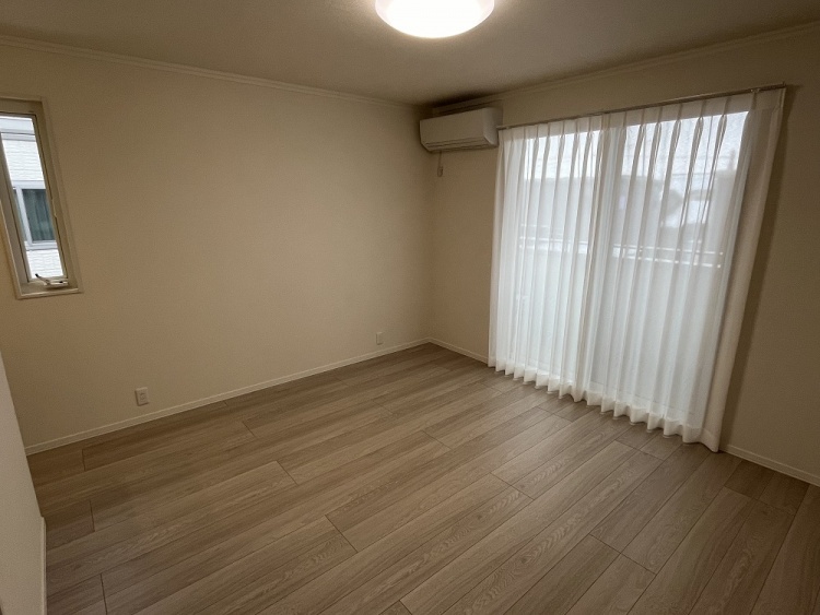 大きな窓から柔らかな光が差し込む落ち着いた雰囲気の部屋。白いカーテンが風に揺れ、木目調の床が温かみを添えている。壁際にはエアコンが設置され、快適な空間を演出している。