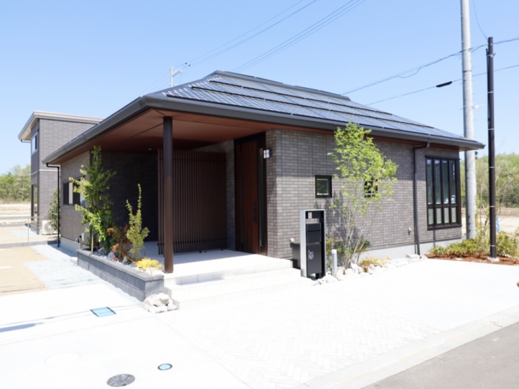 グレーの外壁と黒い屋根を持つ平屋建ての家が、日当たりの良い場所に建っています。玄関には木製の縦格子がアクセントとなり、周りには小さな植栽が植えられています。