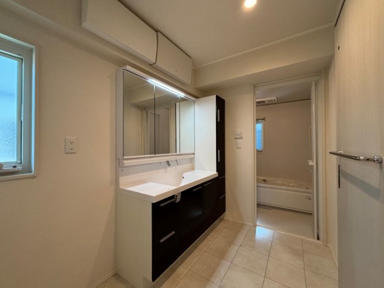 ダークな木目調を基調とした3面鏡の洗面脱衣室。収納や棚、小窓もあります。奥に浴室が見えます。