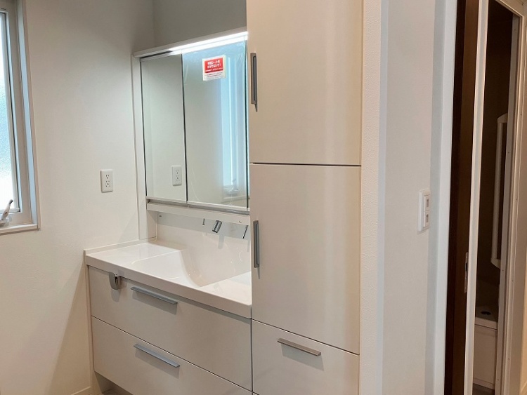 広々とした洗面スペース。白を基調とした洗面台は収納が豊富で、使い勝手が良さそう。大きな鏡が備え付けられ、明るく清潔感のある空間。奥にはキッチンが見え、家事動線が考えられたレイアウトになっています。