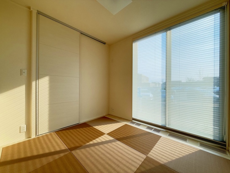 大きな窓から柔らかな陽光が差し込む畳敷きの和室。ブラインド越しに映る光と影が美しく、静かで落ち着いた空間を演出している。淡い色の引き戸が部屋の一角にあり、シンプルながらも温かみのある雰囲気が漂う。