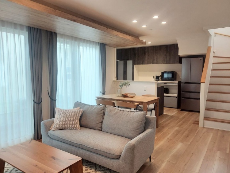 大きな窓からやわらかな光が差し込むリビング。グレーのソファと木製の家具が並び、奥には階段とキッチンが見えます。