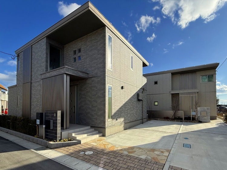 青空の下に建つモダンな住宅。グレーのタイル外壁が特徴的で、シンプルながらも存在感のあるデザイン。広々とした駐車スペースと整えられたエントランスが、落ち着いた街並みに調和している。