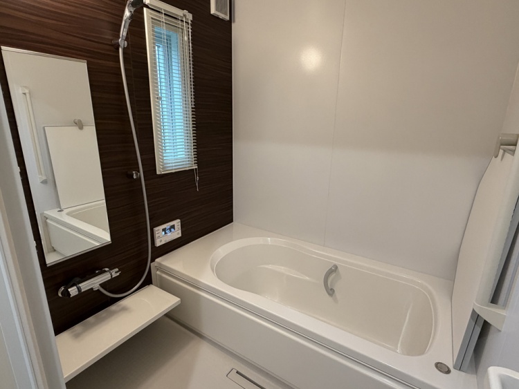 白い浴槽があるバスルームで、壁はダークウッド調のデザインです。窓にはブラインドがかかり、手前にはシャワーと鏡が設置されています。