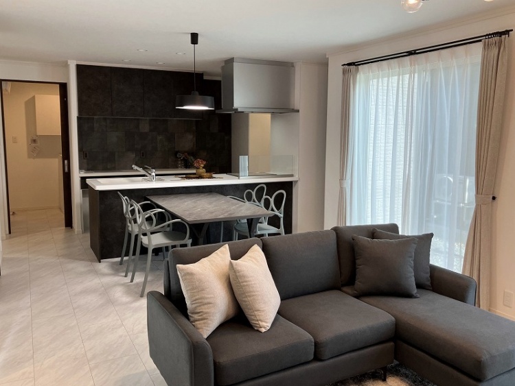 広々としたリビングに、グレーのL字型ソファと丸いコーヒーテーブルが配置されています。ふかふかのラグが床に広がり、くつろぎの空間を演出しています。